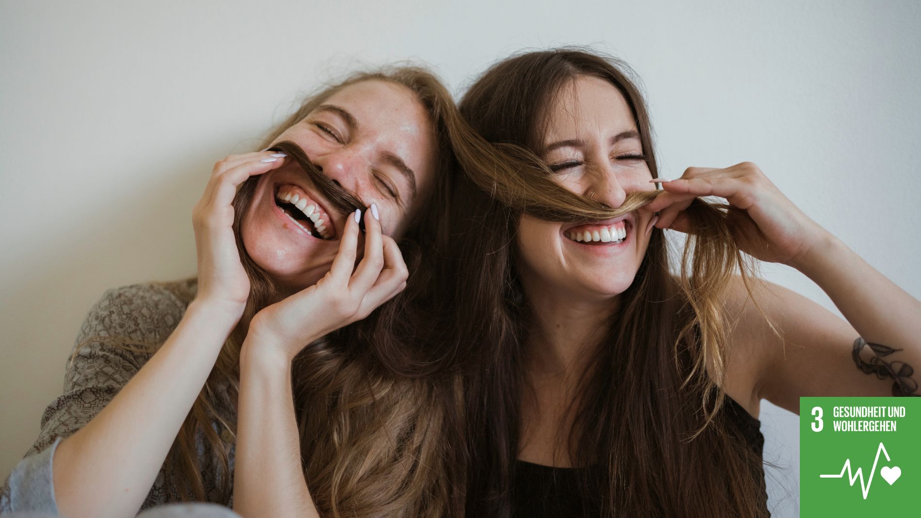 Zwei lachende Frauen formen mit ihren Haaren einen Schnurrbart